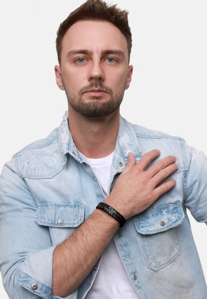 Bracciale in pelle intrecciata nera al polso, indossato con una camicia in denim azzurro chiaro caratterizzata da dettagli logori e bottoni decorativi.