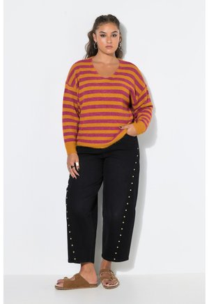 Vrouw draagt een gestreepte oranje en roze trui, zwarte broek met gouden studs aan de zijkanten, en bruine slippers, staand tegen een witte muur.