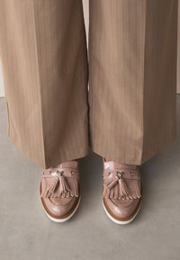Mocassins en cuir verni rose avec détail à franges, semelle compensée marron et quincaillerie argentée, portés avec un pantalon à rayures beige.