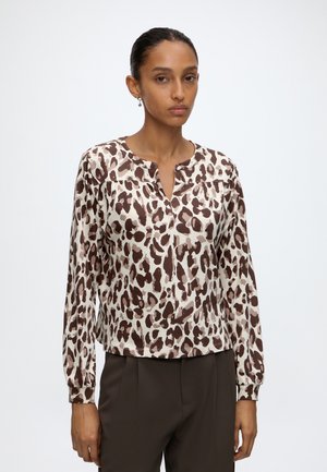 Vrouw draagt een blouse met lange mouwen, bruin-wit luipaardprint, een gespleten halslijn en donkerbruine broek, staand tegen een effen achtergrond.