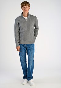 Szary sweter w warkoczowy splot z kołnierzem na pół zamka, w połączeniu z niebieskimi dżinsami i białymi trampkami, na jednolitym białym tle.