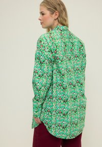 Camicia verde a fantasia con maniche lunghe, caratterizzata da un mix di dettagli rossi e bianchi. Il design è floreale e astratto, con una silhouette ampia.