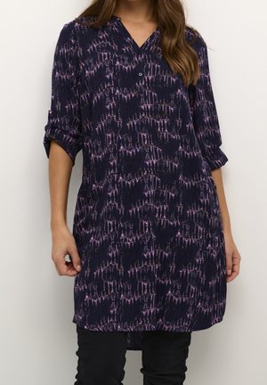 Femme portant une tunique bleu marine aux manches retroussées avec un motif abstrait rose, assortie à un pantalon noir.