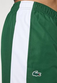 Shorts verzi dintr-un material texturat, având o dungi verticală albă și un mic logo brodat Lacoste în colțul din jos.