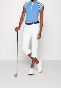 Golfoutfit med en blå kortärmad skjorta med randiga ärmar, vita korta byxor och vita golfskor, som håller en golfklubba.