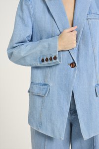Blazer en denim bleu clair avec un col cranté, deux poches avant et des boutons marron. Présente une coupe décontractée et des détails de couture subtils.