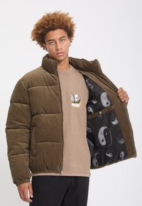 Jeune homme aux cheveux bouclés portant une veste matelassée vert olive montrant une doublure intérieure noire avec des motifs yin-yang et une chemise beige avec un imprimé tête de mort.