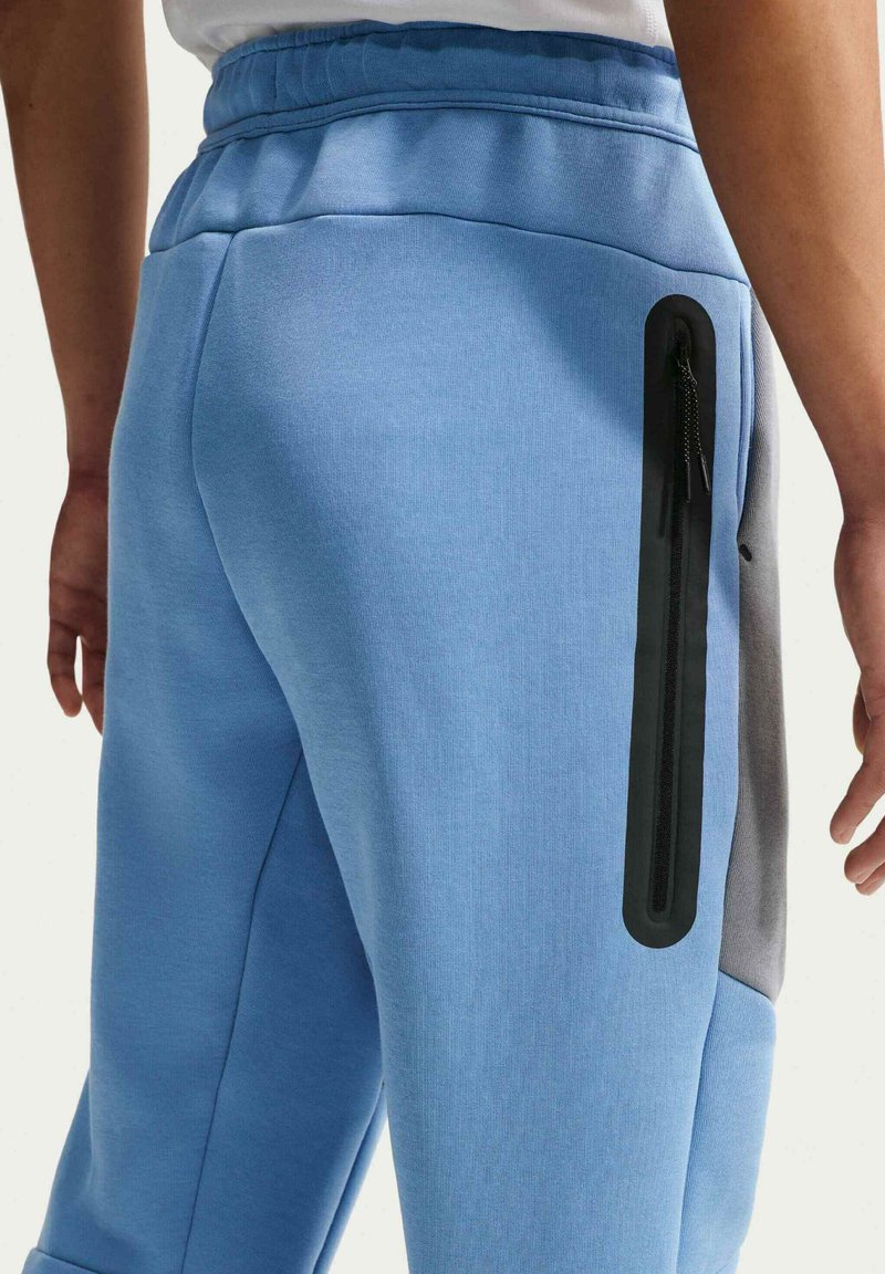 Pantaloni da jogging azzurri realizzati in tessuto morbido, con una tasca nera con cerniera sul fianco e un pannello grigio accentato. Vita elasticizzata visibile.