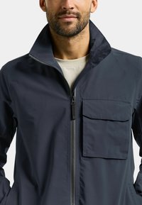 Marineblauwe waterdichte jas met een hoge kraag, een volledige ritssluiting en een borstzak. Soepele textuur met minimale designelementen.