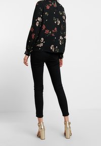 Schwarz-bluse mit floralem Muster und gerafften Ärmeln, kombiniert mit schwarzen Skinny Jeans und goldenen Blockabsatzschuhen. Weißer Hintergrund.