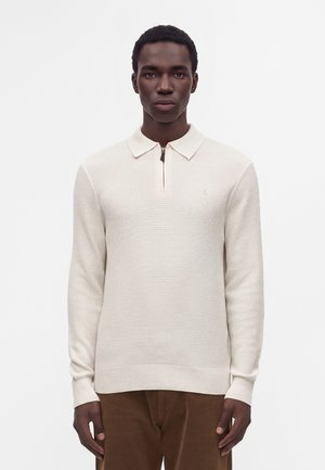 Homme portant un pull en maille crème avec un col demi-fermeture éclair et un pantalon marron, debout devant un fond blanc uni.