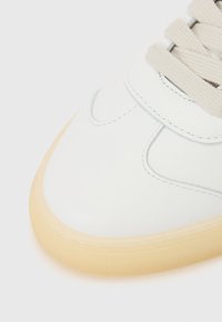 White leather sneaker with a smooth texture, featuring light grey stitching, flat beige laces, and a semi-transparent rubber sole.