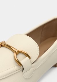 Gros plan sur une chaussure bateau en cuir crème avec un détail en métal doré à l'avant et une doublure intérieure beige visible.