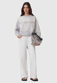 Grijze cropped sweatshirt met "ALLSAINTS" in rhinestones, gecombineerd met witte wijde broek. Doorzichtige zilveren tas met zwarte banden, platte sandalen.