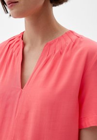 Blouse rose avec un col en V, ornée de plis verticaux au niveau du col et de manches courtes. Le tissu semble léger et lisse.