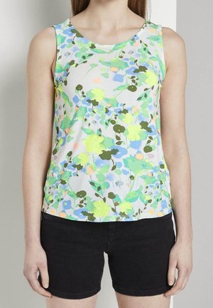 Frau trägt ärmelloses weißes Top mit grünem, blauem und gelbem Blumenmuster kombiniert mit schwarzen Denim-Shorts.