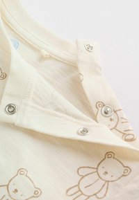Gros plan sur un vêtement pour bébé couleur crème avec des boutons-pression et des imprimés d'ours en peluche marron sur un tissu doux.