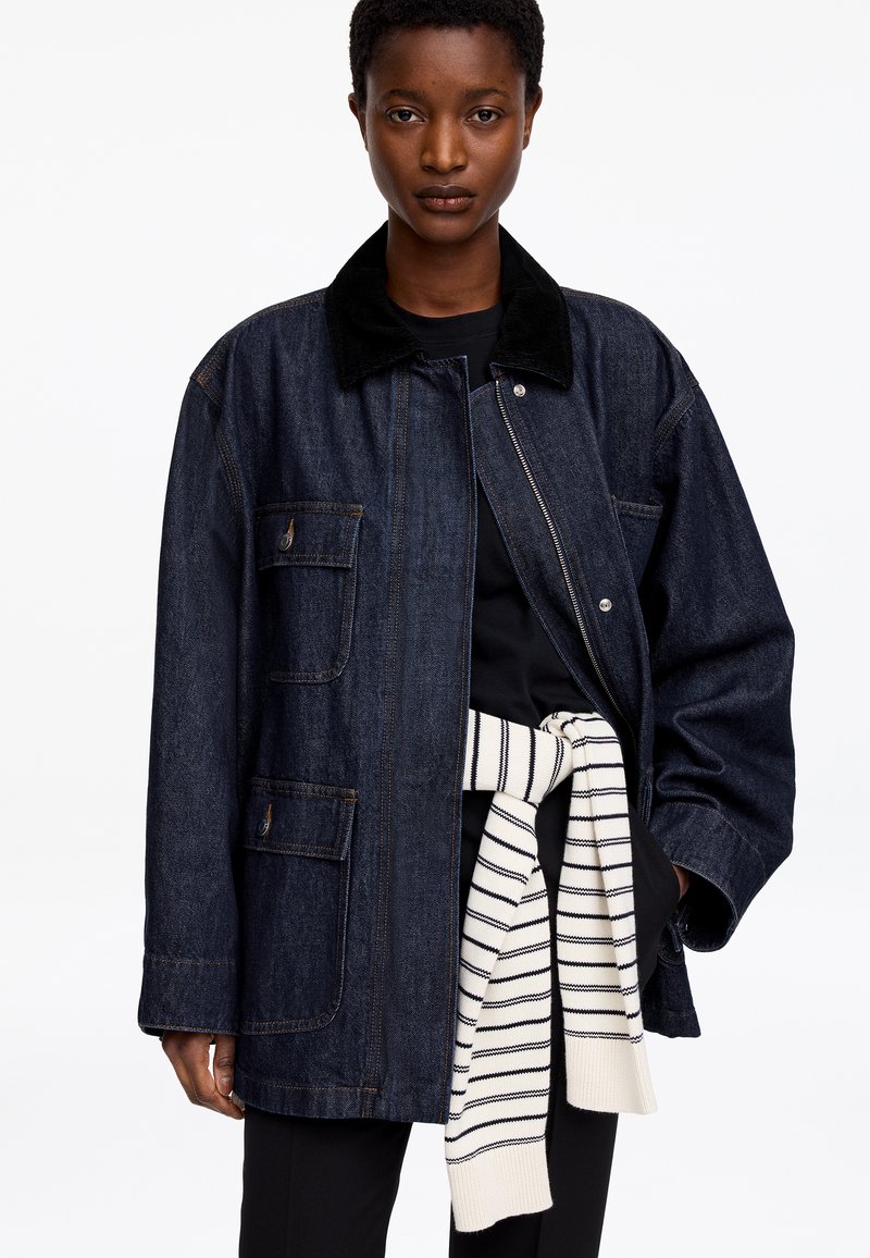 Personne portant une veste en jean sombre oversize avec de grandes poches et une fermeture éclair, chemise noire, et pull rayé noué autour de la taille.