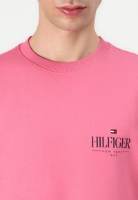 Rosa hettegenser laget av bomull, med en rund ribbestrikket halslinning. Inkluderer en svart logo med "HILFIGER" tekst og liten flaggdetalj.