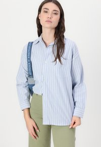 Camicia azzurra a righe con maniche lunghe e tasca frontale, abbinata a pantaloni verdi oliva. Un accessorio con una tracolla blu pende dalla spalla.