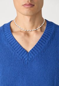 Pull bleu en V avec texture côtelée et col, porté avec un collier en perles composé de perles lisses et texturées alternées.