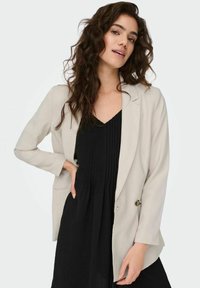 Lichtgrijze blazer met een gladde textuur, enkele knoopsluiting en twee voorkezen. Draagt over een zwarte geplooide jurk.