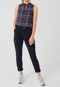 Donna che indossa un top a maniche senza spalline verde e viola a quadri, pantaloni cargo neri arrotolati e sneakers platform argentate, in piedi con le mani in tasca.