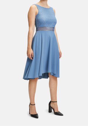 Kobieta w niebieskiej sukience midi bez rękawów z koronki i szyfonu z asymetrycznym dołem oraz czarnych szpilkach z paskiem na kostce, stojąca na białym tle.