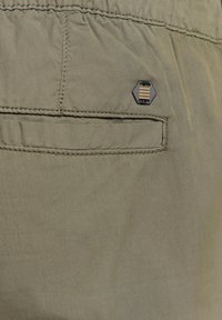 Khaki korte broek met een zachte textuur, voorzien van een achterzak en een zwarte hexagonale label met gouden lijnen, gestikte details langs de naden.