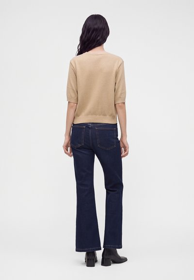 Femme aux longs cheveux bruns portant un pull beige à manches courtes, un jean bleu foncé et des bottines noires, debout dos à l'objectif sur un fond uni.