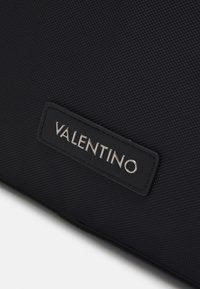 Närbild av svart texturerad tyg med en sydd rektangulär svart etikett som har silverfärgade "VALENTINO"-bokstäver.