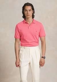 Jauns vīrietis ar vidēja garuma matiem, tērpies rozā polo kreklā, kas iešūts augsta vidukļa baltos pleitētos biksēs, ar pulksteni uz kreisā plaukstas.