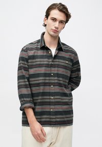 Młody mężczyzna ubrany w ciemnoszary, czerwony i beżowy w paski koszulę z podwiniętymi rękawami oraz jasne spodnie, stojący na jednolitym tle.