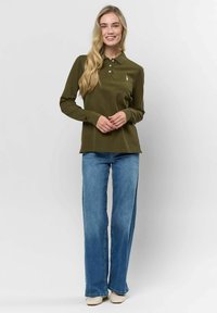 Hosszú ujjú olíva zöld polo felső, három gombos nyakkal és hímzett logóval, világoskék bővített lábú farmerrel és semleges színű cipővel párosítva.