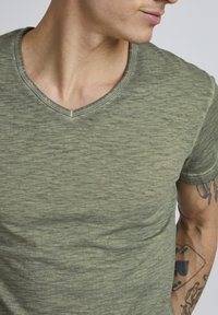 Maslinasto zeleni T-shirt s V-izrezom od teksturiranog materijala, s kratkim rukavima i suptilnim varijacijama boje. Minimalistički dizajn bez uzoraka.