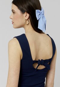 Débardeur bleu marine à côtes avec un dos en scoop présentant un détail en nœud papillon. Les cheveux du modèle sont ornés d'un ruban rayé bleu et blanc. Boucles d'oreilles en or en forme de créole.