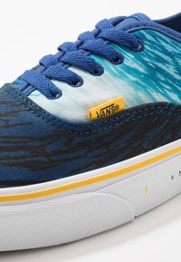 Sneaker in tela blu con motivo ondulato, etichetta gialla del marchio Vans, suola in gomma bianca e dettagli cuciti sulla suola intermedia.