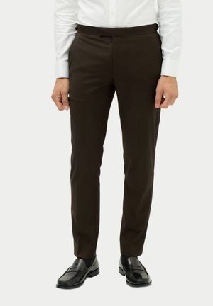 Man wearing slim fit dark brown dress pants, black leather loafers, and a white buttoned shirt standing against a plain background.