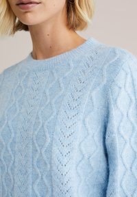 Pulover din lână de culoare bleu deschis, tricotat, realizat dintr-un material moale și texturat. Dispune de un guler rotund și de modele complicate de puncte în diagonală.