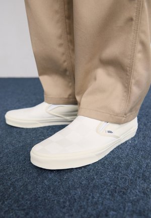 Des chaussures blanches à enfiler avec un motif en damier, fabriquées dans un matériau lisse. Elles possèdent une semelle en caoutchouc et des accents de marque sur le côté.