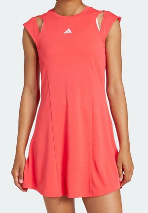 Femme portant une robe sans manches corail rose Adidas avec un petit logo blanc sur la poitrine et des détails découpés aux épaules.