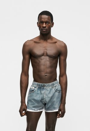 Atletický tmavší muž s krátkymi vlasmi stojaci pred jednoduchým pozadím, oblečený v svetlomodrých riflových kraťasoch s elastickým pásom.