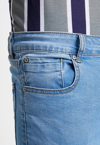 Jeans de mezclilla azul claro con un diseño estándar de cinco bolsillos y costuras en naranja contrastante. Presentan un pequeño bolsillo para monedas y herrajes metálicos.