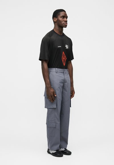 Μαύρο κοντομάνικο t-shirt με κόκκινο σχέδιο και γκρι παντελόνια cargo. Τα παντελόνια έχουν πλαϊνές τσέπες και άνετη εφαρμογή. Μαύρα παπούτσια με λευκές ρίγες.