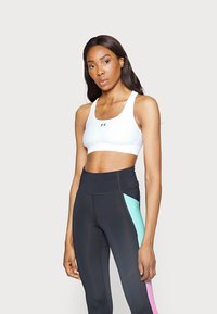 Witte sportbh met brede banden en logo, gecombineerd met zwarte hoogwijde leggings met turquoise en roze zijpanelen. Zachte textuur.