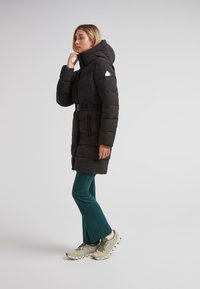 Kvinde justerer kraven på en lang sort pufferjakke med bælte, iført tealfarvede bukser og beige sneakers, stående mod en ensartet lys baggrund.