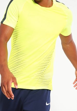 Man wearing bright yellow sports shirt with black dot pattern and navy blue Nike shorts in motion against white background.