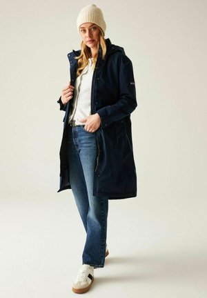 Kobieta w beżowej dzianinowej czapce, granatowym długim płaszczu, białym swetrze, niebieskich dżinsach i białych trampkach z czarnymi i beżowymi akcentami.