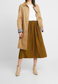 Trench beige con puños de cuadros en blanco y negro, combinado con una falda amarilla con pliegues y botas de tobillo beige. Blusa blanca con puntos dorados.