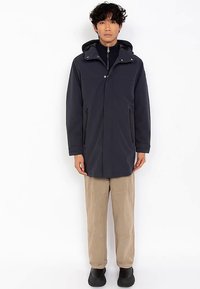 Giacca impermeabile blu navy con cappuccio, chiusura frontale con cerniera e tasche laterali, indossata sopra pantaloni beige e scarpe nere. Tessuto liscio e opaco.